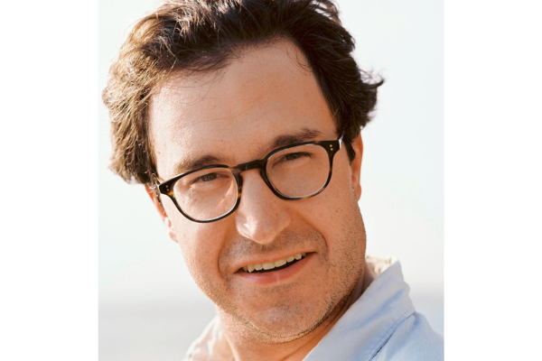 A man wearing glasses and a blue shirt, smiling while standing against a neutral background