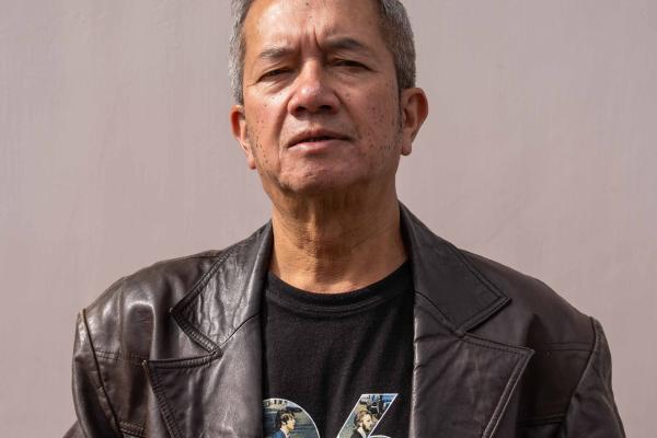 An individual wearing a leather jacket and a printed t-shirt stands with a neutral expression against a plain background.