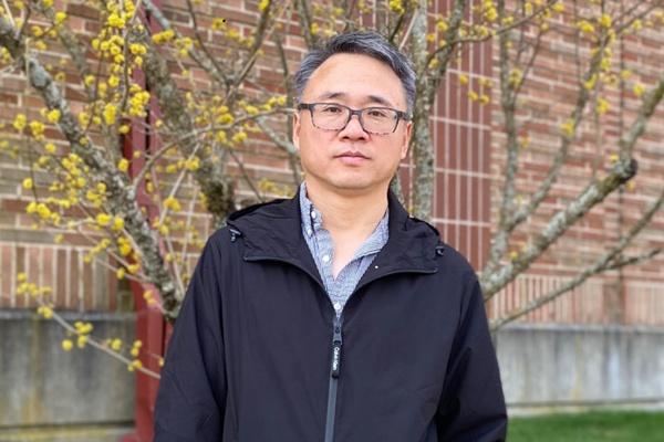 A man wearing glasses and a black jacket stands outdoors in front of a brick wall and a tree with yellow buds.