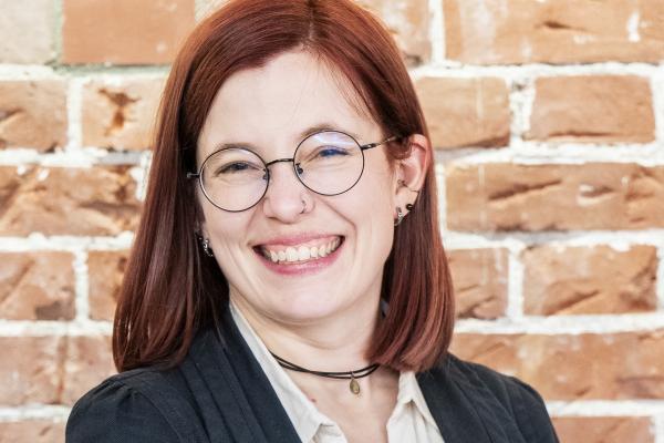 A woman with red hair and glasses smiles brightly against a rustic brick wall backdrop
