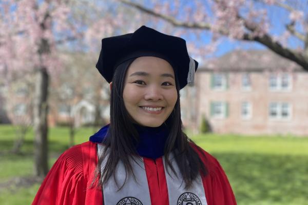 A woman dressed in a graduation gown and cap stands against a backdrop of a tall tree.