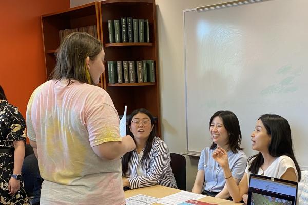 A person stands and talks to three seated individuals behind a table at an indoor event. The table displays name tags, papers, and a laptop. Other people are present in the background.