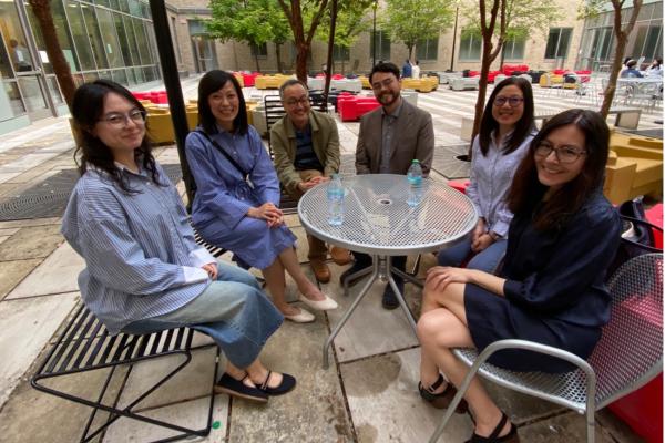 Six people sit around a circular metal table outdoors, smiling at the camera. The table has three water bottles, and the setting includes trees and modern buildings.