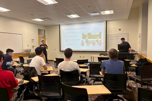  A classroom with students seated at desks facing two presenters; a slide about elementary school is projected on a screen at the front of the room. 