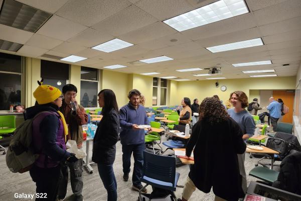  A group of people stand and talk in a classroom with green chairs and overhead lights. Some hold plates of food. The room appears to be set up for a casual gathering or event. 