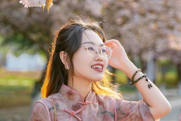 A woman wearing glasses and a floral dress stands in front of a tree, smiling and enjoying the outdoors.  