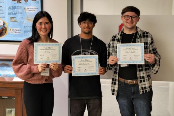Three students pose with their certificates in front of a wall, smiling with pride