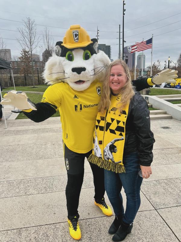 A woman (Meridith Linch) stands smiling with a mascot in front of a cityscape.