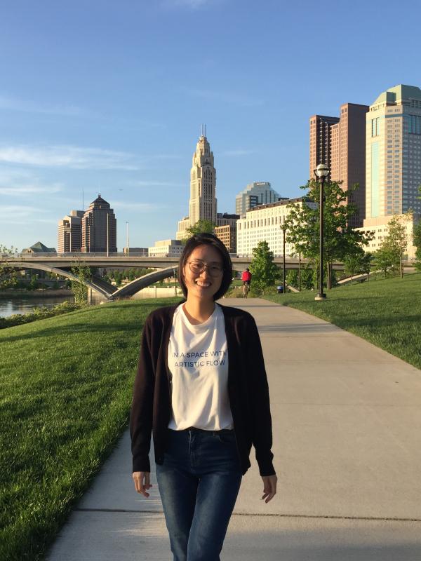 Woman walking on a pathway in a park with skyscrapers and a bridge in the background under a clear blue sky