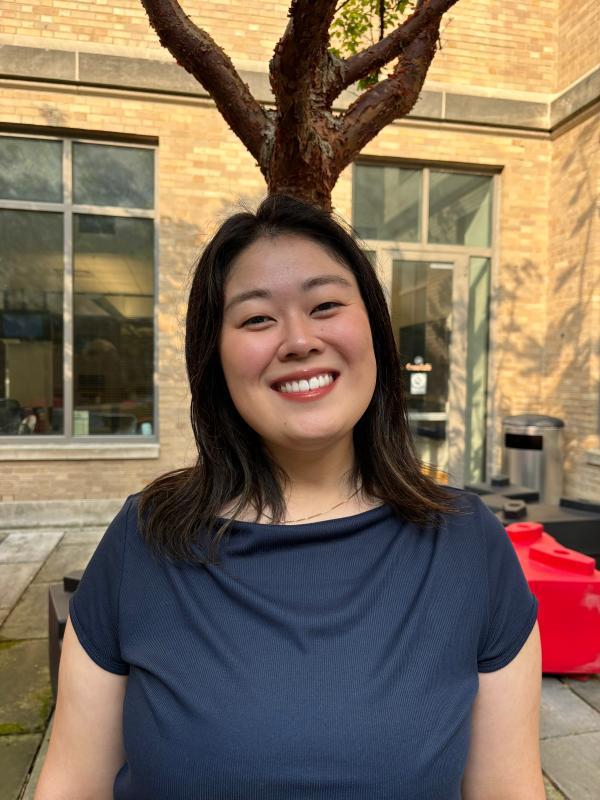 A woman in a navy blue top is standing outdoors in front of a brick building. She is smiling, and a tree with bare branches is visible behind her.