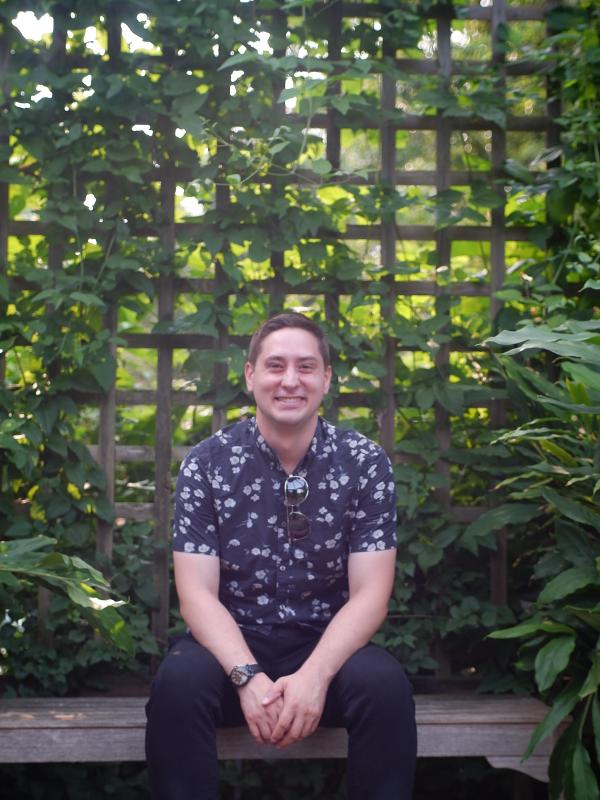 Smiling man sitting at a bench with foliage behind him. 