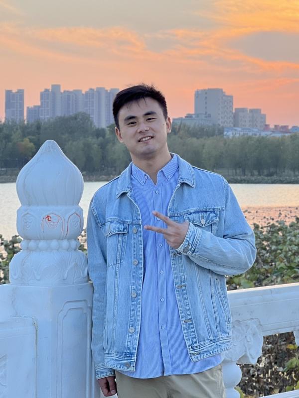 A man stands on a bridge with a city skyline and sunset in the background, making a peace sign