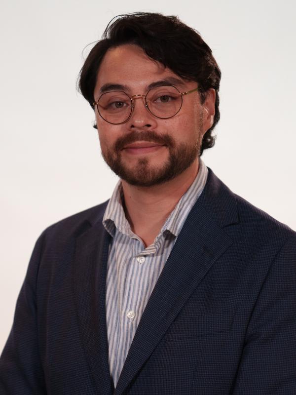 A man with glasses and a beard is wearing a dark blazer over a striped shirt, standing against a plain, light-colored background.