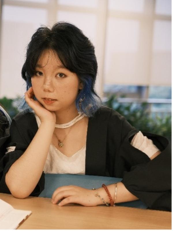 A woman with blue hair is seated at a table, looking thoughtfully at her surroundings.  