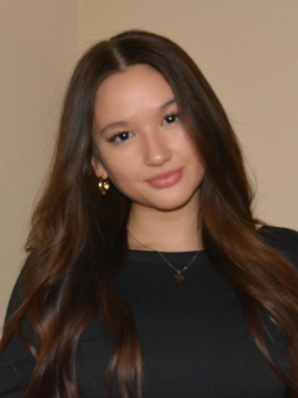  A woman with long brown hair wearing a black top and gold earrings stands in front of a plain beige background. 