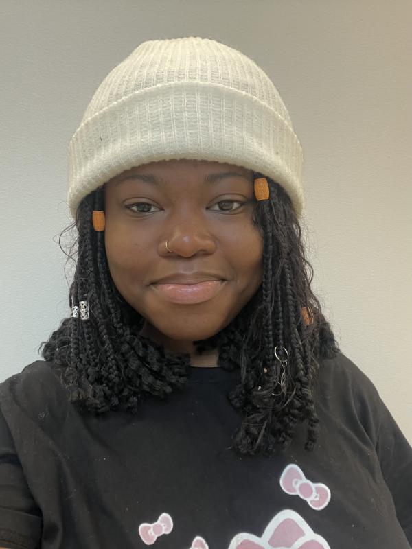 A smiling woman with black braided hair, wearing a white knit beanie and a black t-shirt with a pink graphic, stands against a plain light background.