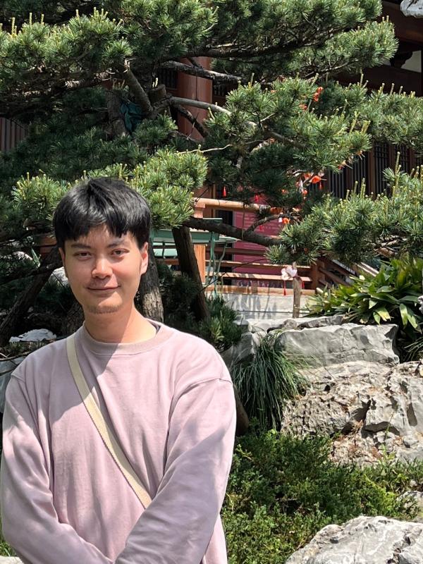  A man in a light pink sweatshirt stands in front of rocks, greenery, and a tree, with a building partially visible in the background. 