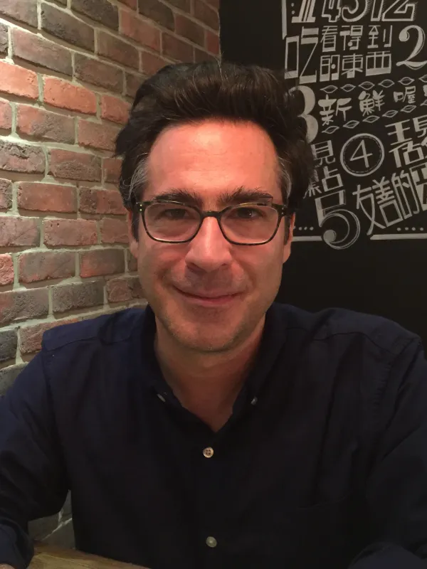 Man with dark hair and glasses with a dark collared shirt sititng next to a brick wall and smiling