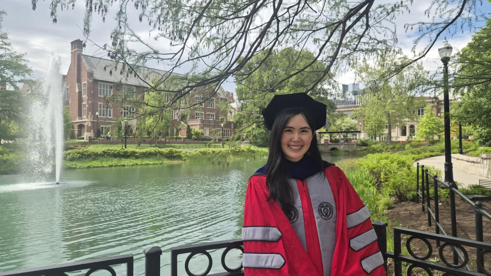 Person in a graduation gown by a lake with a fountain and historic buildings in the background.