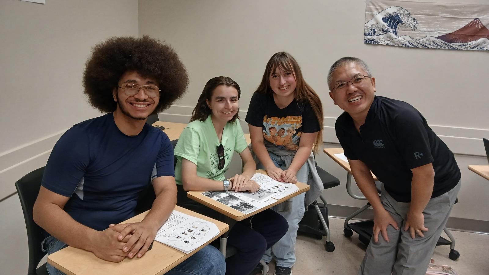 3 students sitting at desks with instructor leaning over, all posed and smiling for picture
