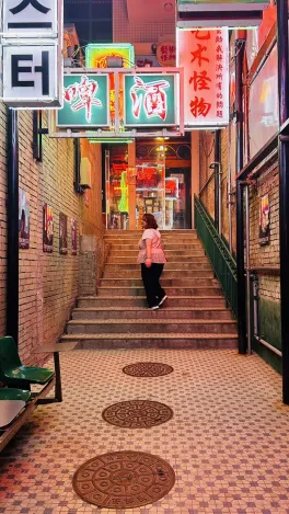 A woman walks up a dimly lit alley stairway, surrounded by glowing neon signs with Korean characters. The scene feels vibrant and urban.