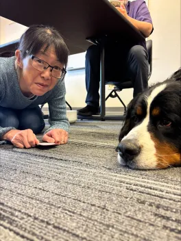 A woman crouches on a carpeted floor beside a large, relaxed dog. She holds a card and smiles slightly. The setting is a casual indoor space.