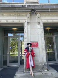 Woman in a graduation gown stands in front of Hagerty Hall, holding a diploma, with two glass doors and stone columns in the background.